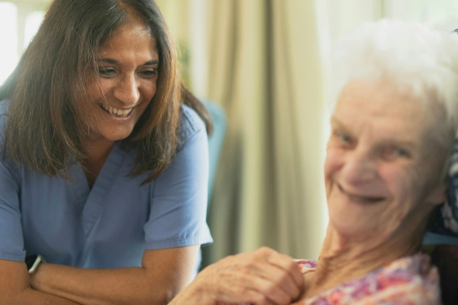 A caregiver in blue scrubs smiles with an older woman.