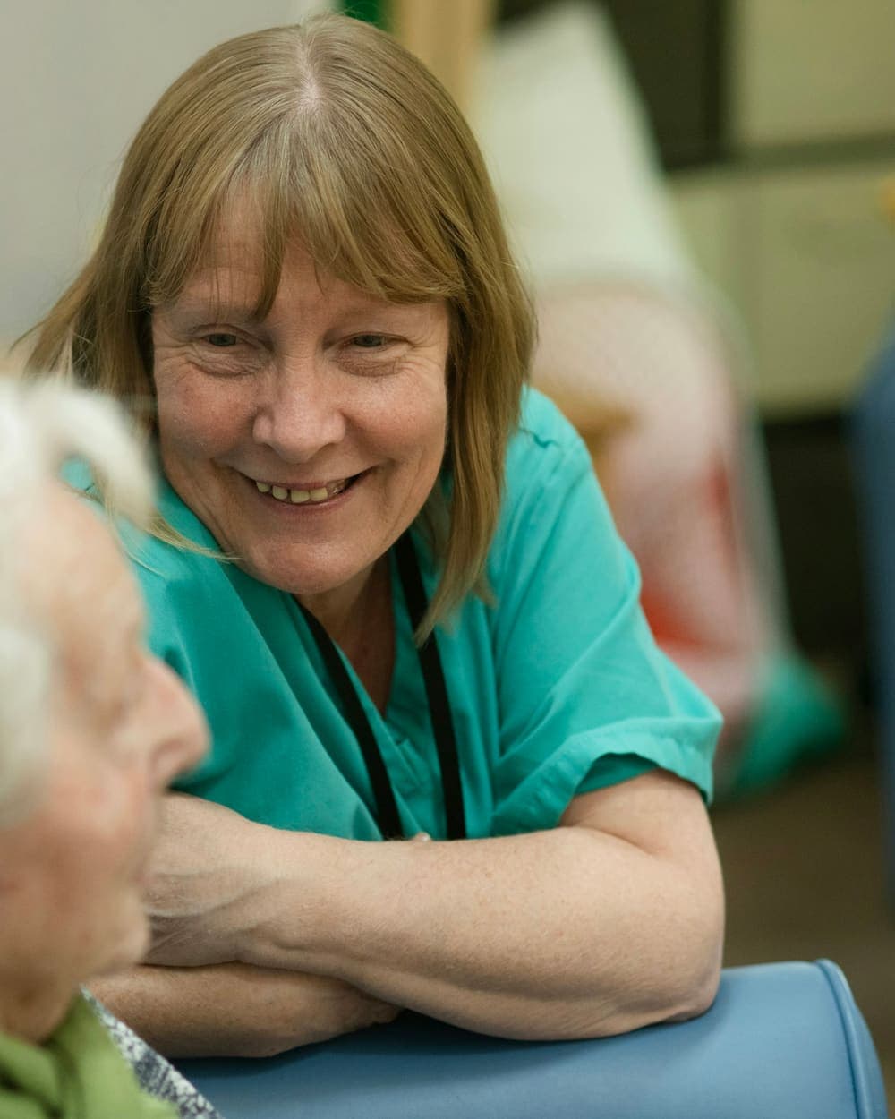 A smiling caregiver in teal scrubs chats with an older woman.