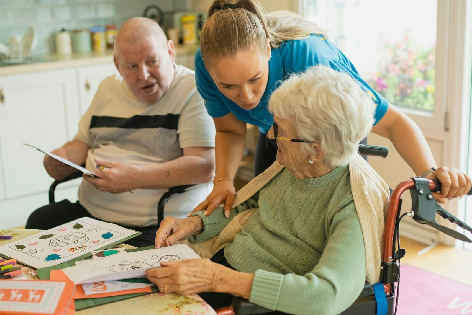 A caregiver helps an older woman with a coloring page at a kitchen table as an older man looks on.