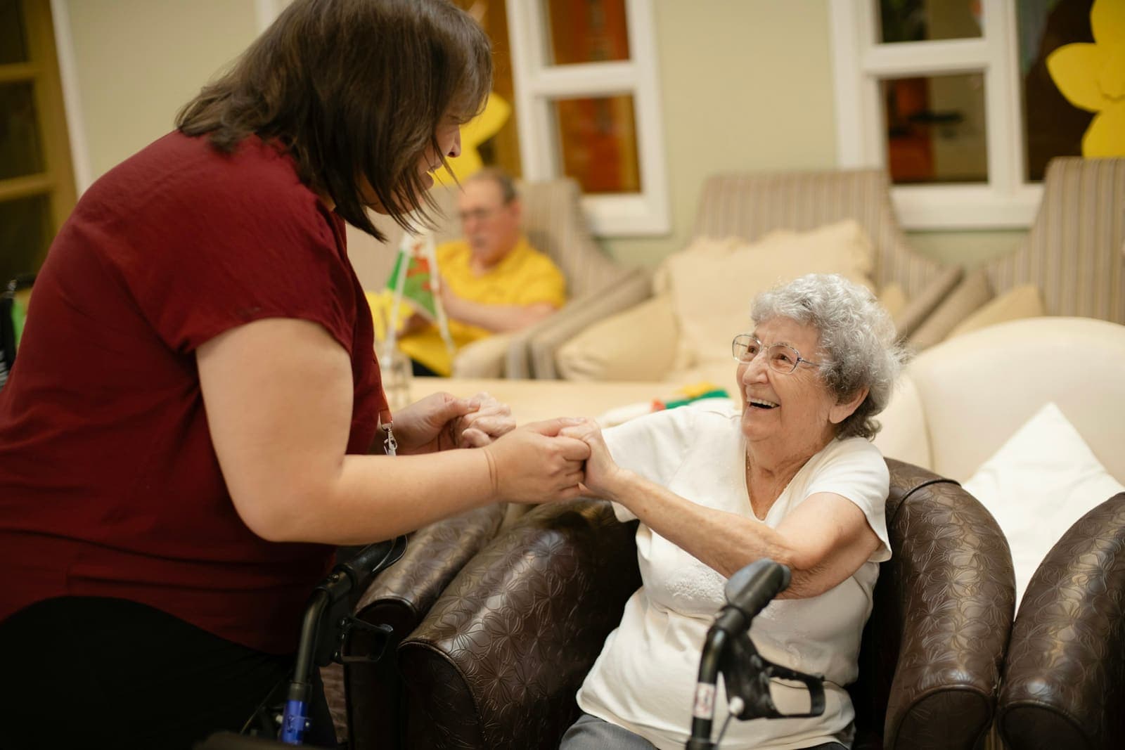 A caregiver holds an older woman’s hands as she rises from her armchair, with her walker beside her.