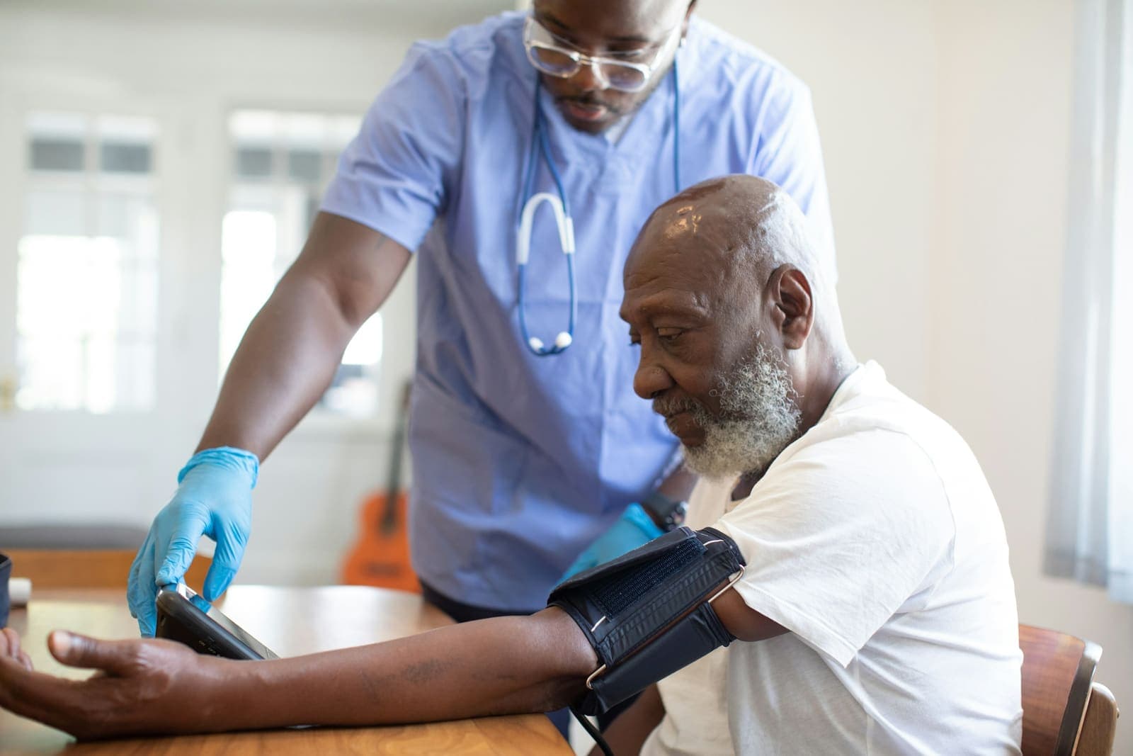 A nurse in blue scrubs checks an older man’s blood pressure at a table.